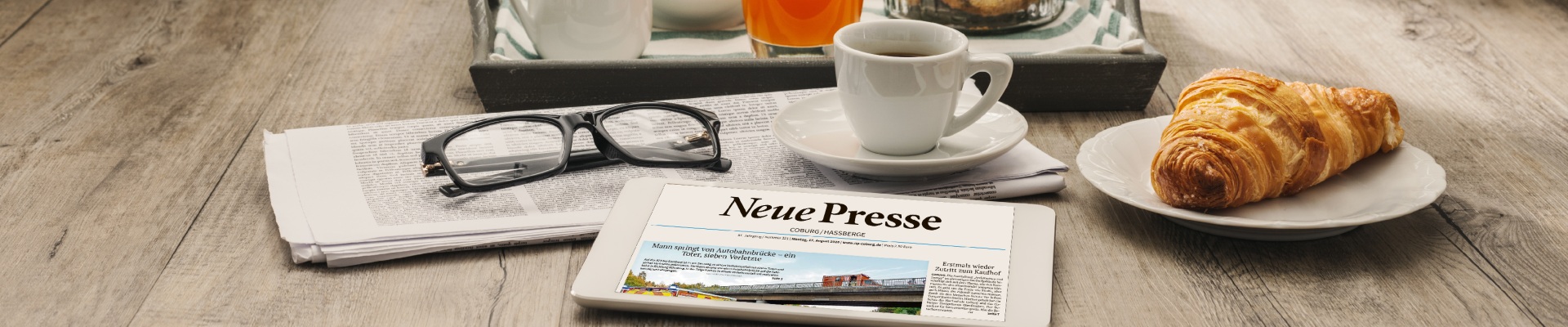 Tablet shows a news article on a wooden table next to a cup of coffee, a croissant on a plate, and reading glasses laid over a folded newspaper tray of drinks behind them.